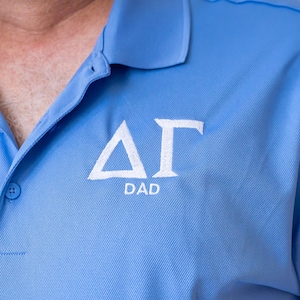 May include: Light blue polo shirt with white embroidered Greek letters and the word "DAD" underneath. The shirt has a button-down collar and a textured fabric.