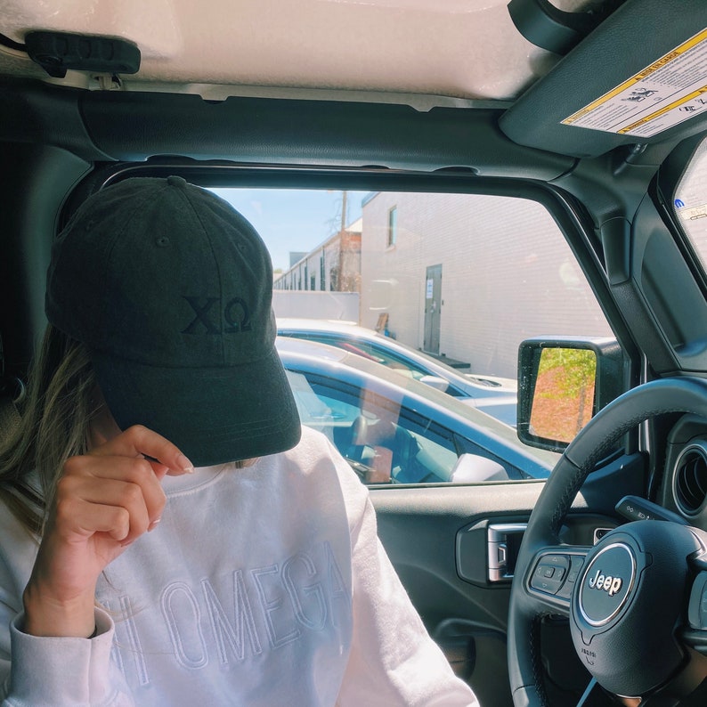 May include: A person wearing a black baseball cap with the letters "XO" embroidered on the front, sitting in the driver's seat of a black Jeep. The person is wearing a white sweatshirt with the word "OMEGA" written on the front.