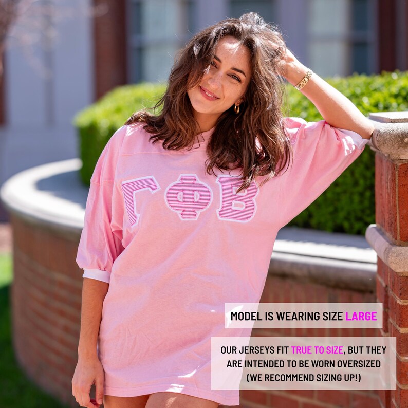 May include: A light pink oversized jersey featuring white and pink striped Greek letters on the chest. The garment has short sleeves with white cuffs and a crew neckline. Text on the image states: MODEL IS WEARING SIZE LARGE. OUR JERSEYS FIT TRUE TO SIZE, BUT THEY ARE INTENDED TO BE WORN OVERSIZED (WE RECOMMEND SIZING UP!).