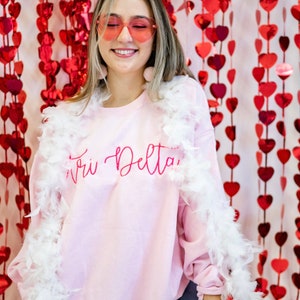 May include: A pink sweatshirt with the text "Tri Delta" in pink script. The sweatshirt is worn by a person with blonde hair and pink sunglasses. The person is standing in front of a red heart-shaped backdrop.