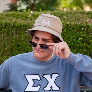 May include: A light blue sweatshirt with a white embroidered "EX" on the front. The person is wearing a beige bucket hat with a white embroidered "EX" on the front and black sunglasses.