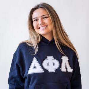 Pu&ograve; includere: Felpa con cappuccio blu navy con lettere greche bianche e grigie. La felpa ha una tasca frontale e un cappuccio con coulisse. Le lettere sono cucite sul davanti. La persona nell'immagine sta sorridendo.