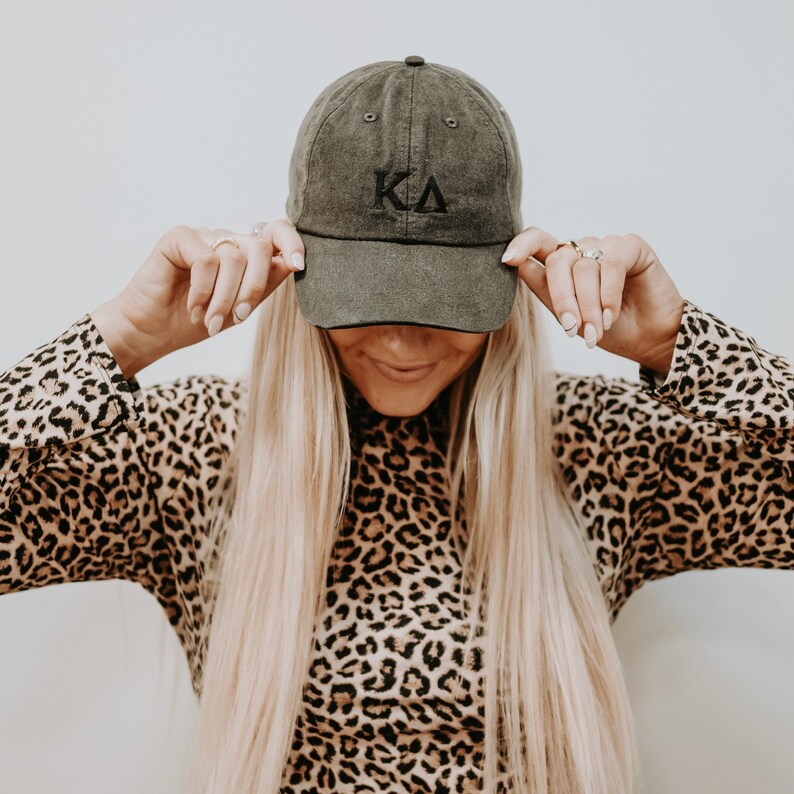 May include: A woman wearing a leopard print shirt is holding a dark green baseball cap with the letters "KA" embroidered on the front.