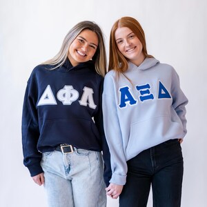 Pu&ograve; includere: Due persone indossano felpe con cappuccio abbinate. Una &egrave; blu navy con lettere greche bianche, l'altra &egrave; azzurra con lettere greche blu. Entrambe le felpe hanno una tasca frontale e un cappuccio con coulisse.