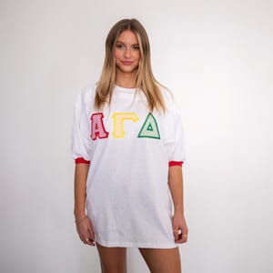 May include: A white short-sleeved t-shirt featuring the Greek letters Alpha, Gamma, and Delta in red, yellow, and green embroidered appliqu&eacute; on the chest. The shirt has red trim on the sleeve cuffs and a crew neckline, providing a classic athletic look.