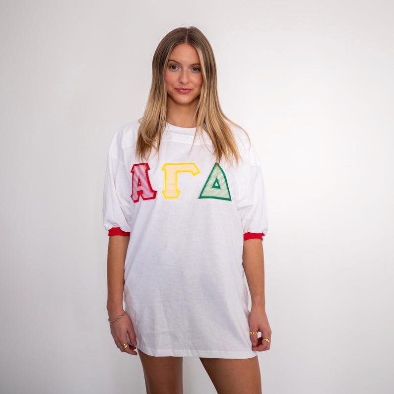 May include: A white short-sleeved t-shirt featuring the Greek letters Alpha, Gamma, and Delta in red, yellow, and green embroidered appliqu&eacute; on the chest. The shirt has red trim on the sleeve cuffs and a crew neckline, providing a classic athletic look.