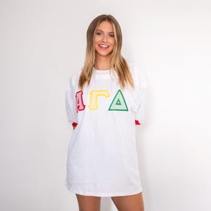 May include: A white short-sleeved t-shirt featuring the Greek letters Alpha, Gamma, and Delta in red, yellow, and green respectively. The shirt has a crew neckline and red trim on the sleeve cuffs, with a loose, comfortable fit and a plain white back.