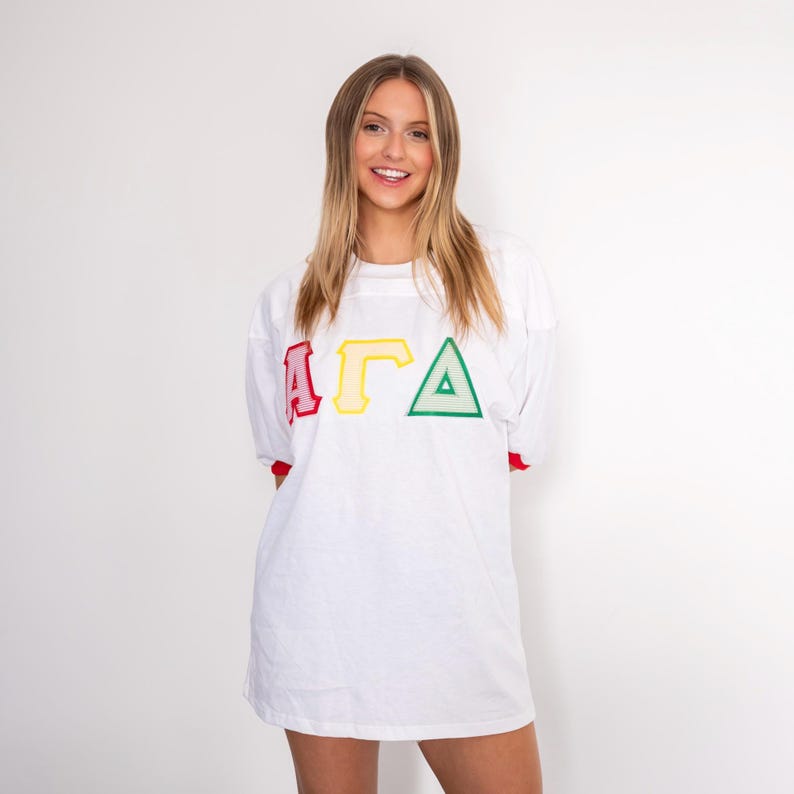 May include: A white short-sleeved t-shirt featuring the Greek letters Alpha, Gamma, and Delta in red, yellow, and green respectively. The shirt has a crew neckline and red trim on the sleeve cuffs, with a loose, comfortable fit and a plain white back.