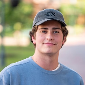May include: A young man with short brown hair is wearing a blue sweatshirt and a gray baseball cap with the letters "ΣΧ" embroidered on the front.