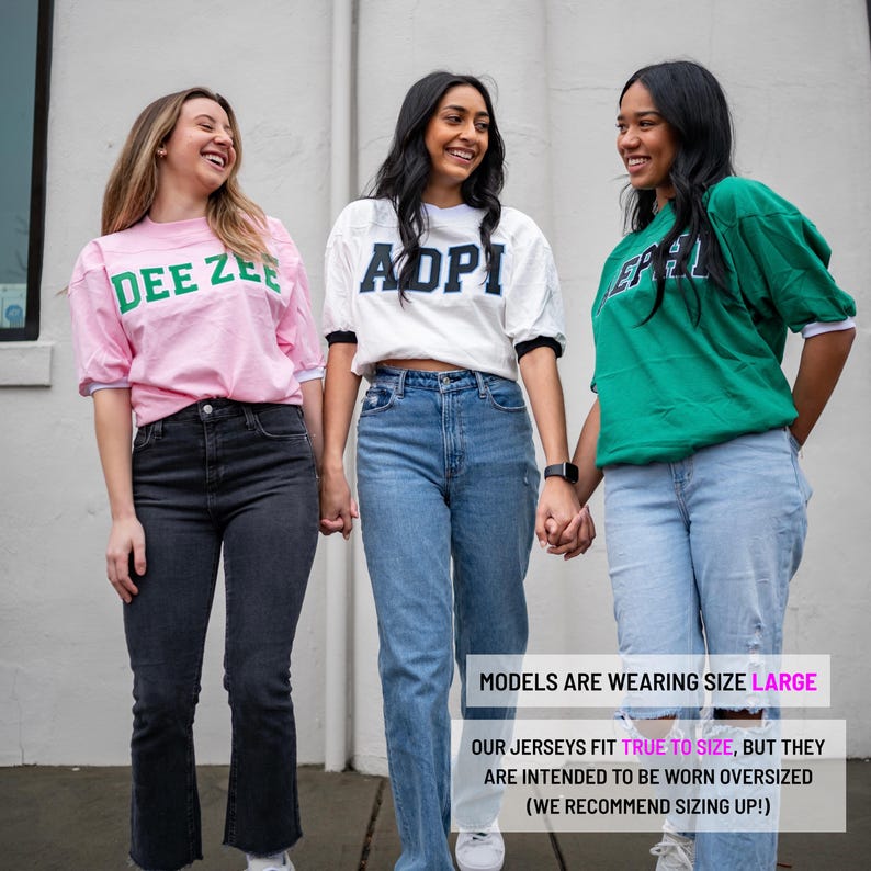 May include: Three short-sleeved jerseys in pink, white, and green with block lettering reading DEE ZEE, ADPI, and AEPHI. The jerseys feature contrasting sleeve cuffs and are paired with various denim jeans. Text on image: MODELS ARE WEARING SIZE LARGE. OUR JERSEYS FIT TRUE TO SIZE, BUT THEY ARE INTENDED TO BE WORN OVERSIZED (WE RECOMMEND SIZING UP!)
