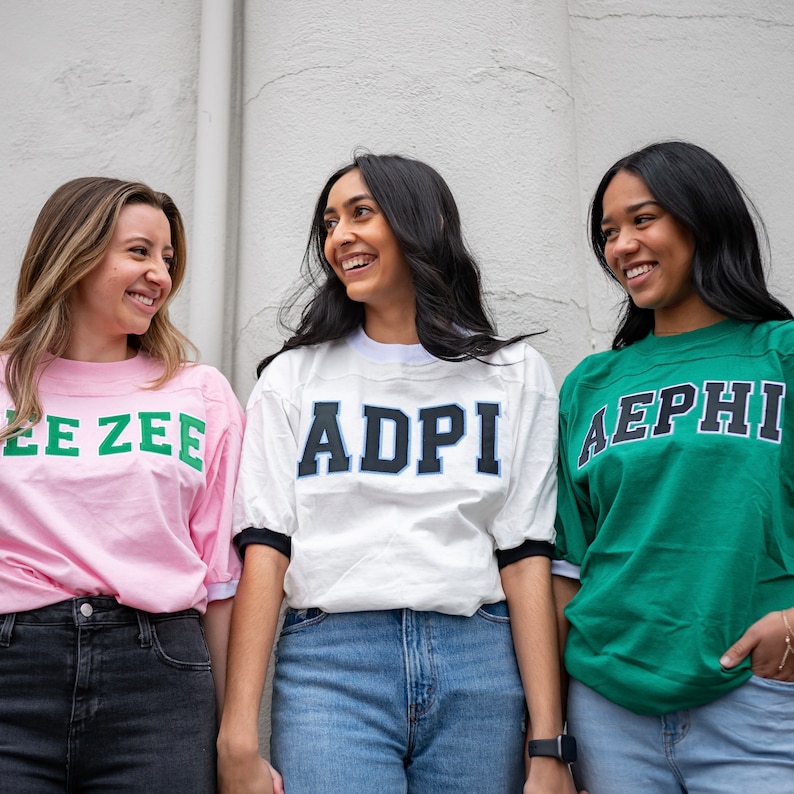 May include: Three short-sleeved crewneck t-shirts. The left is light pink with green text reading ZEE ZEE. The center is white with black and blue text reading ADPI. The right is green with dark blue and white text reading AEPHI.