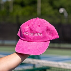 May include: A pink cotton baseball cap with white embroidered text that reads "Zeta Tau Alpha".