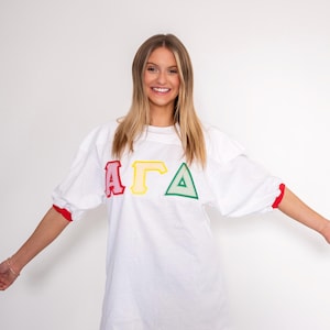 May include: A white oversized jersey shirt featuring red, yellow, and green Greek letters across the chest. The shirt has elbow-length sleeves with red cuffs and a crew neckline. The letters are outlined in a bold, striped pattern.