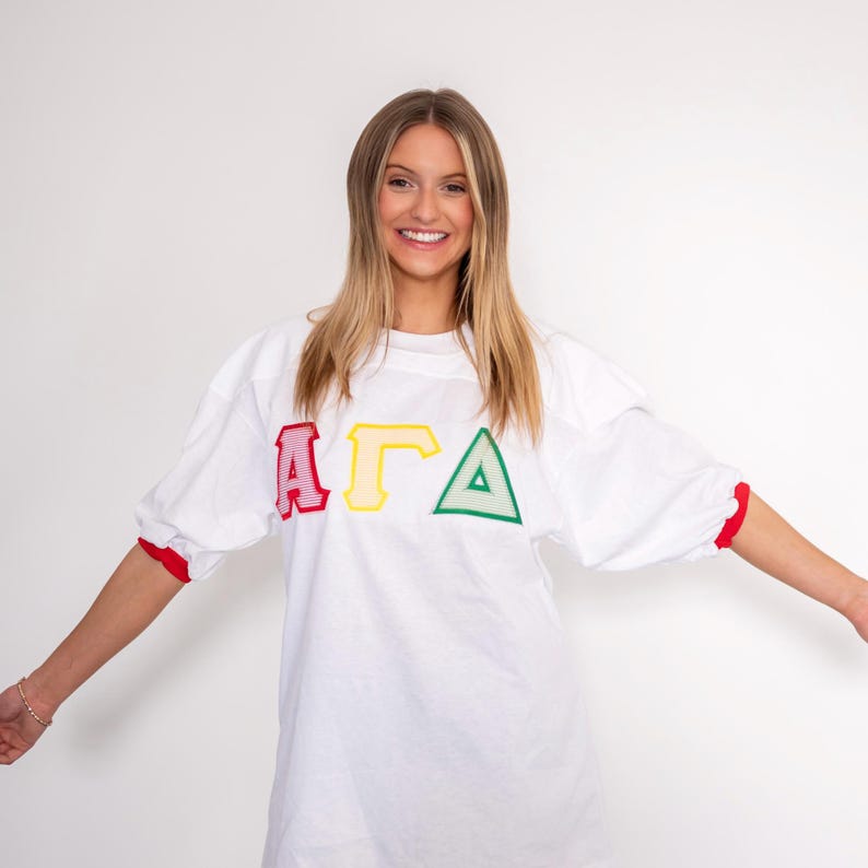 May include: A white oversized jersey shirt featuring red, yellow, and green Greek letters across the chest. The shirt has elbow-length sleeves with red cuffs and a crew neckline. The letters are outlined in a bold, striped pattern.