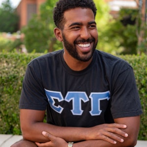 May include: A man wearing a black t-shirt with a blue and white "ΣΤΓ" logo. He is smiling and has a beard.