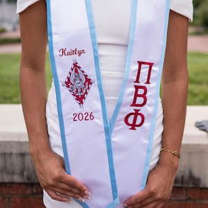 May include: White graduation sash with light blue trim. The sash features the name "Kaitlyn" in red script, a crest, and the year "2026". The Greek letters "ΠΒΦ" are also displayed in red.