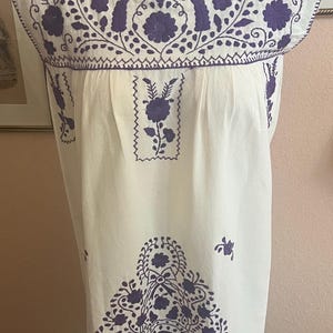 May include: A cream-colored sleeveless dress featuring dark purple floral embroidery. The neckline and chest area are adorned with intricate botanical patterns, while a large floral basket motif is centered on the lower front of the garment.