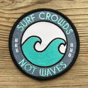 以下が含まれることがあります： 「Surf Crowds Not Waves」というテキストとターコイズブルーの波のデザインが施された、青と白の刺繍パッチです。