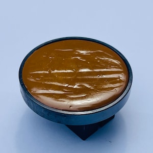 May include: A round, brown,  wax stamp with a black plastic base. The wax is smooth and shiny.