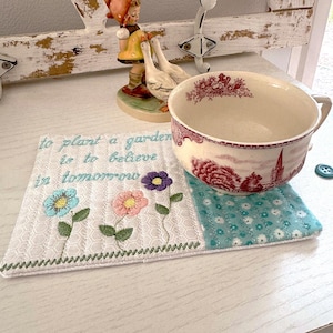May include: A floral embroidered coaster with the text "to plant a garden is to believe in tomorrow" supports a large teacup. The teacup has a red and white landscape design. Decorative buttons and figurines are in the background.