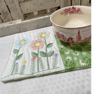 以下が含まれることがあります： 青、ピンク、黄色の刺繍の花が施された、花柄のキルトプレースマット。 赤と白の風景デザインの白いボウルがプレースマットの上にあります。 プレースマットには、緑色の花柄のセクションがあります。