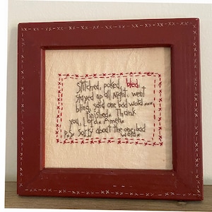 Op de afbeelding: Een ingelijst stuk borduurwerk met rode steken op een crème achtergrond. De borduurwerktekst luidt: "Genaaid, geprikt, gebloed. De hele nacht wakker gebleven, blind geworden, een slecht woord gezegd. Klaar. Dank je, Lorax. Amelie. P.S. Sorry voor het ene slechte woord."