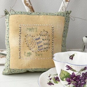May include: A small, square, green and tan pillow with the words "Merry Eggs Day" embroidered on it. The pillow is sitting on a white surface next to a white teacup and saucer with a purple flower design.