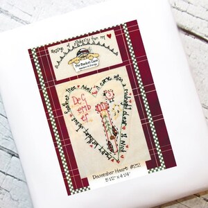Op de afbeelding: Een rode en witte geruite achtergrond met een hartvormig ontwerp. Het hart is gevuld met een witte achtergrond en een handgestikt ontwerp van een kerstsok, een kerstboom en de woorden "December Heart #212 5 1/2" x 4 1/4"