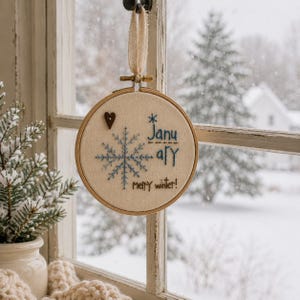 May include: A circular wooden embroidery hoop containing off-white fabric with blue stitched snowflake embroidery, a small brown heart button, and the blue embroidered text "January" above the brown stitched text "merry winter!" with a ribbon.