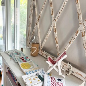 Pu&ograve; includere: Una cornice in legno bianco anticato con un motivo a rombi &egrave; montata su un muro. Una bottiglia di vetro trasparente avvolta in iuta con un design a cuore &egrave; appoggiata su una mensola bianca, insieme a oggetti trapuntati e una sedia a sdraio in miniatura.