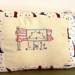 May include: A white pillow with a red, white, and blue embroidered flag and the word "Liberty" on it. The pillow has a blue and white fabric with stars on it.