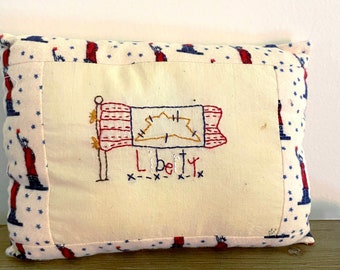 May include: A white pillow with a red, white, and blue embroidered flag and the word "Liberty" on it. The pillow has a blue and white fabric with stars on it.