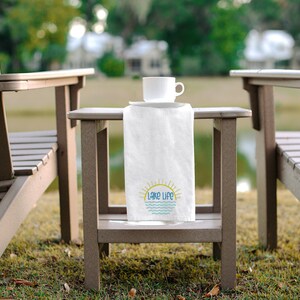 May include: A white rectangular kitchen towel draped over a small brown side table. The towel features a graphic of a yellow sun rising over blue wavy lines with the text Lake Life printed in blue letters. A white ceramic mug sits on the table.