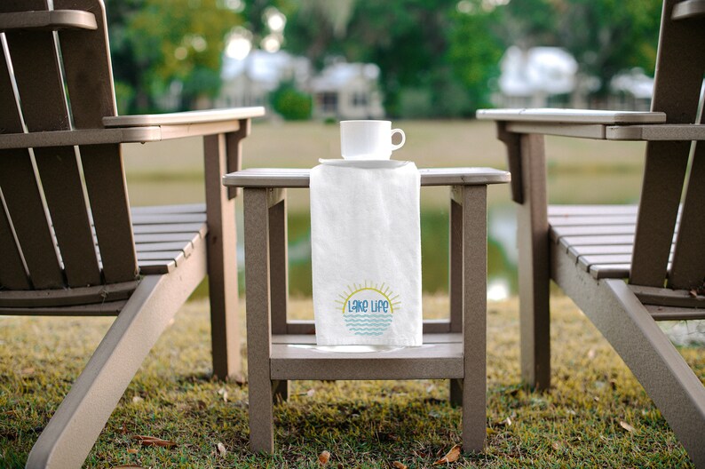 May include: A white rectangular kitchen towel draped over a small brown side table. The towel features a graphic of a yellow sun rising over blue wavy lines with the text Lake Life printed in blue letters. A white ceramic mug sits on the table.