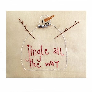 May include: White fabric with an embroidered snowman design. The snowman has a carrot nose, black eyes, and a red heart. The snowman is holding two branches and has jingle bells around its neck. The text "jingle all the way" is embroidered in red below the snowman.