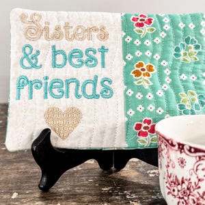 May include: A decorative quilted plaque with the words "Sisters & best friends" embroidered in teal and tan. The plaque has a heart and floral pattern, and is displayed on a black stand. A floral teacup is in the foreground.