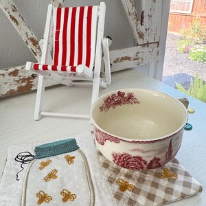 Pu&ograve; includere: Una piccola sedia a sdraio in legno bianco con tessuto a righe rosse e bianche. Una ciotola in ceramica con un motivo floreale rosso poggia su un sottobicchiere trapuntato. &Egrave; presente anche un sottobicchiere a forma di barattolo con api ricamate.
