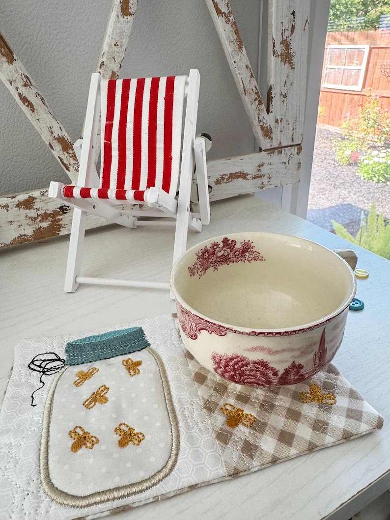 Pu&ograve; includere: Una piccola sedia a sdraio in legno bianco con tessuto a righe rosse e bianche. Una ciotola in ceramica con un motivo floreale rosso poggia su un sottobicchiere trapuntato. &Egrave; presente anche un sottobicchiere a forma di barattolo con api ricamate.