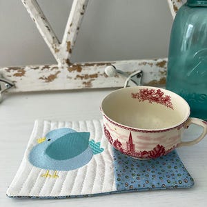 May include: A white quilted coaster with a blue bird design and a blue and white patterned border. The coaster is sitting on a white surface next to a teacup and a glass jar.