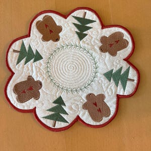 May include: A white, quilted, floral-shaped placemat with a red scalloped border. The placemat features brown bear and green tree appliqués. A green wreath and spiral design are in the center. The placemat is likely for the holiday season.