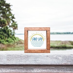 May include: A square wooden sign with a dark brown frame and a white center panel. The center features a graphic of a yellow sun rising over blue wavy lines, with the text "Lake Life" printed in blue letters centered between the sun and the waves.