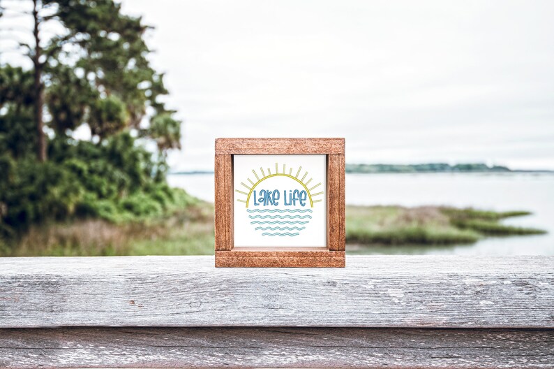 May include: A square wooden sign with a dark brown frame and a white center panel. The center features a graphic of a yellow sun rising over blue wavy lines, with the text "Lake Life" printed in blue letters centered between the sun and the waves.