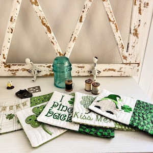 May include: A collection of St. Patrick's Day themed quilted coasters. The coasters feature shamrocks, the text "Luck", "I Pine Back", and "Kiss Me". Other items include a blue glass insulator, spools of thread, and decorative accents.