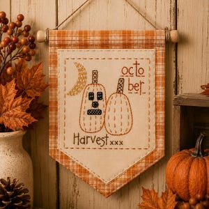 May include: A hanging fabric banner with an orange and white plaid border and a cream-colored center panel. The panel features embroidered designs of two pumpkins, a crescent moon, and the text "octo ber" and "Harvest xxx" in brown thread.