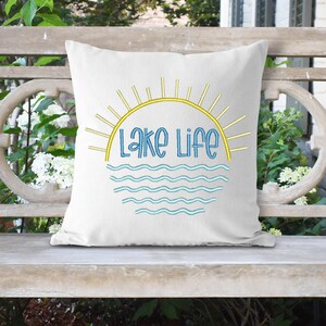 May include: A square white throw pillow featuring a graphic design of a yellow sun rising over blue wavy lines. The text "Lake Life" is printed in blue letters in the center of the design, positioned between the sun rays and the water waves.