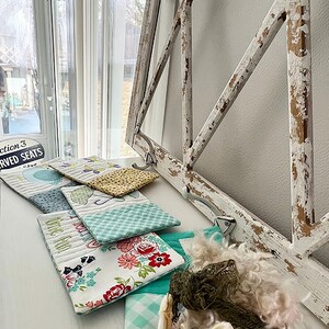 May include: A collection of five small quilted fabric pouches with various floral and geometric patterns. The pouches are arranged on a white surface with a distressed white wooden frame in the background.