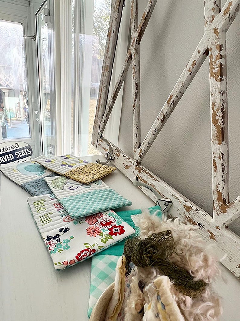 May include: A collection of five small quilted fabric pouches with various floral and geometric patterns. The pouches are arranged on a white surface with a distressed white wooden frame in the background.