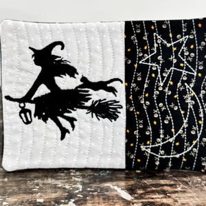 May include: A rectangular quilted fabric coaster featuring a split design. The left side is light gray with white dots and a black silhouette of a witch on a broomstick with a cat. The right side is black with small skull icons and yellow stars.