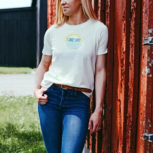 May include: A short-sleeved white t-shirt featuring a graphic on the chest with the text "LAKE LIFE" in blue, positioned below a yellow sun icon and above stylized blue wavy lines representing water, paired with dark blue denim jeans and a brown belt.