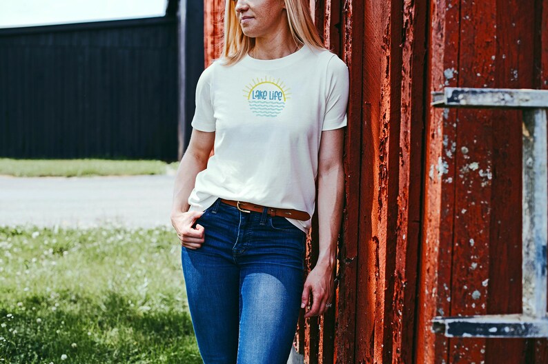 May include: A short-sleeved white t-shirt featuring a graphic on the chest with the text "LAKE LIFE" in blue, positioned below a yellow sun icon and above stylized blue wavy lines representing water, paired with dark blue denim jeans and a brown belt.